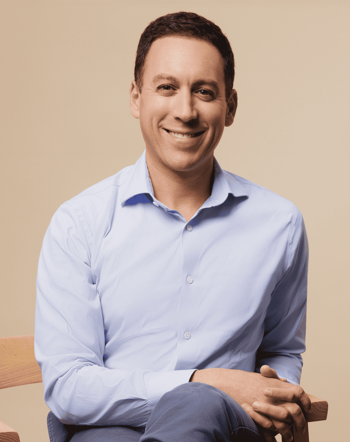 A man in a light blue button-up shirt sits on a wooden chair, smiling with his hands resting on his knee against a beige background.