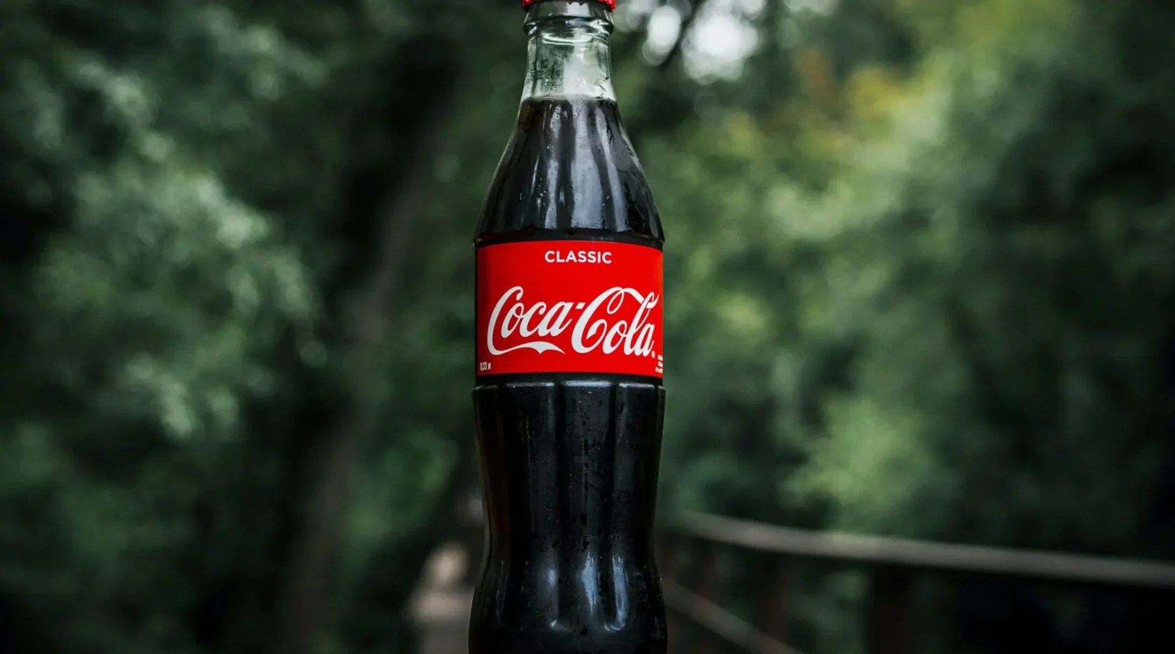 A classic glass bottle of Coca-Cola with a red label is shown outdoors against a blurred background.