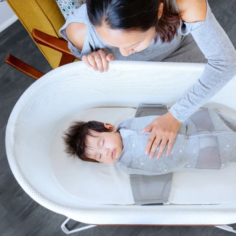 An adult leans over a baby sleeping in a white bassinet gently resting a hand on the baby’s chest. The baby is swaddled in a gray wrap.