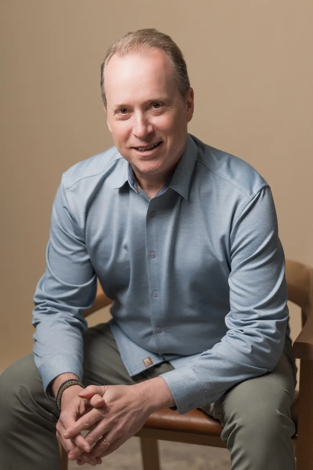 Peter Saperstone, a man with short light hair, is sitting on a chair wearing a light blue button-up shirt and gray pants, smiling at the camera.