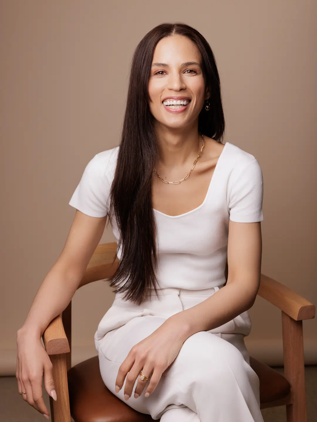Shandra McManus, a woman with long brown hair, dressed in a white top and pants, sits on a wooden chair and smiles at the camera against a neutral beige background.