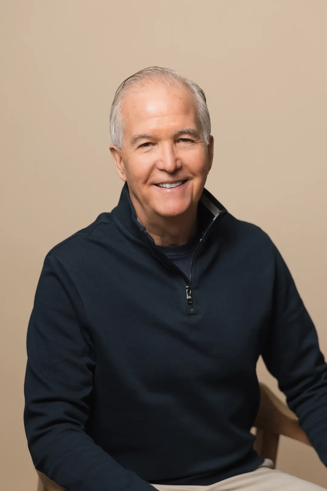 Jim Moffatt, a man with short gray hair, wearing a dark zip-up sweater, sits and smiles in front of a plain beige background.