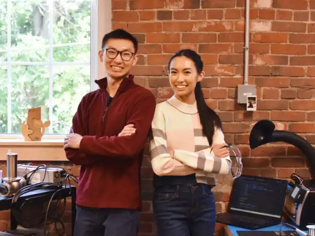 Two people stand with arms crossed and smile in a workspace with exposed brick walls, electronics, a window, and a laptop on a desk.