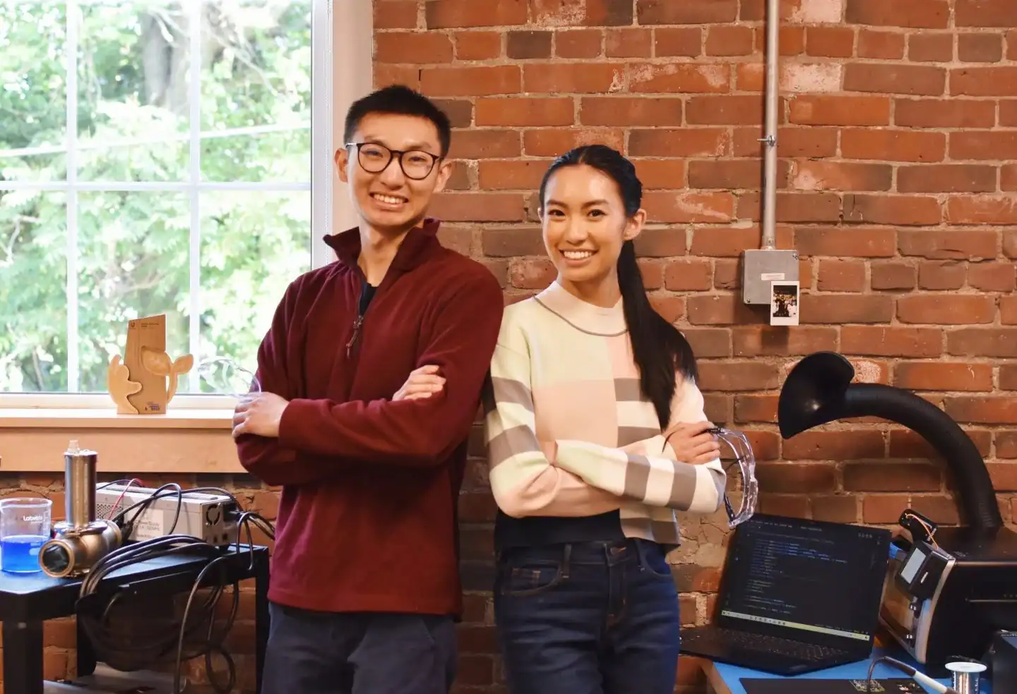 Two people stand with arms crossed and smile in a workspace with exposed brick walls, electronics, a window, and a laptop on a desk.