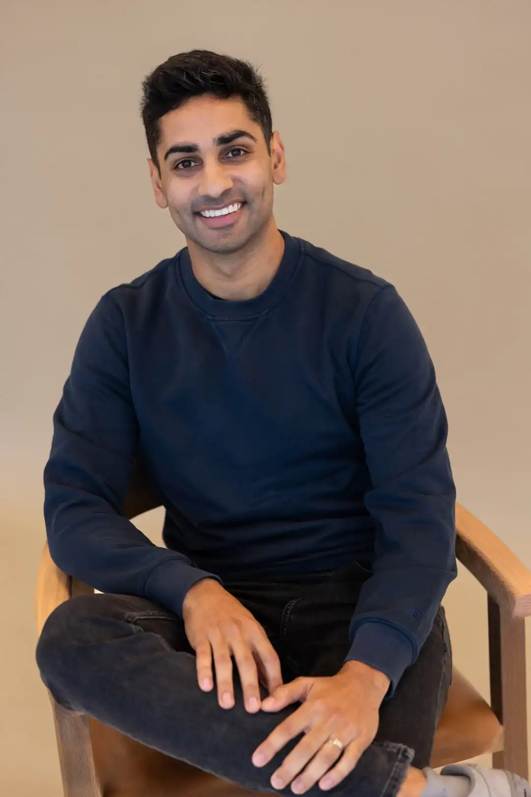Chiraag Deora, a man with dark hair, wearing a navy sweatshirt and black jeans, sits on a wooden chair, smiling with one leg crossed over the other against a plain background.