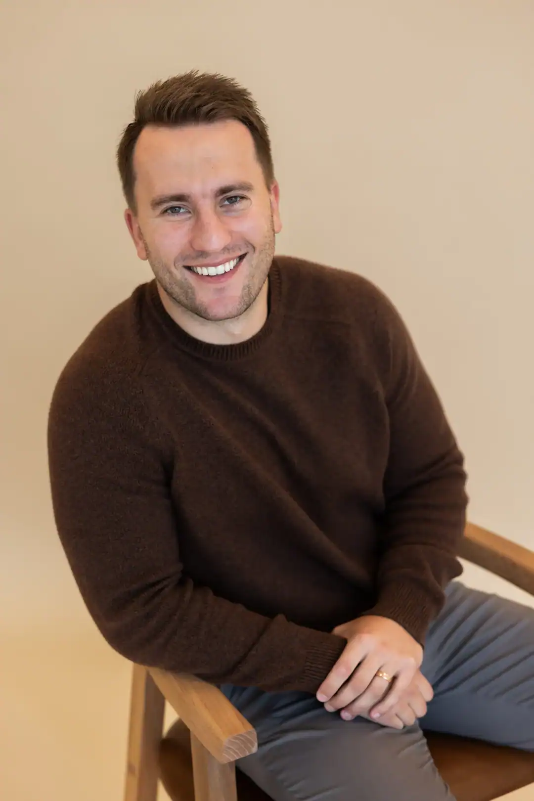 Cameron McGinn, a man with short brown hair, wearing a brown sweater and gray pants, sits on a wooden chair and smiles at the camera against a beige background.