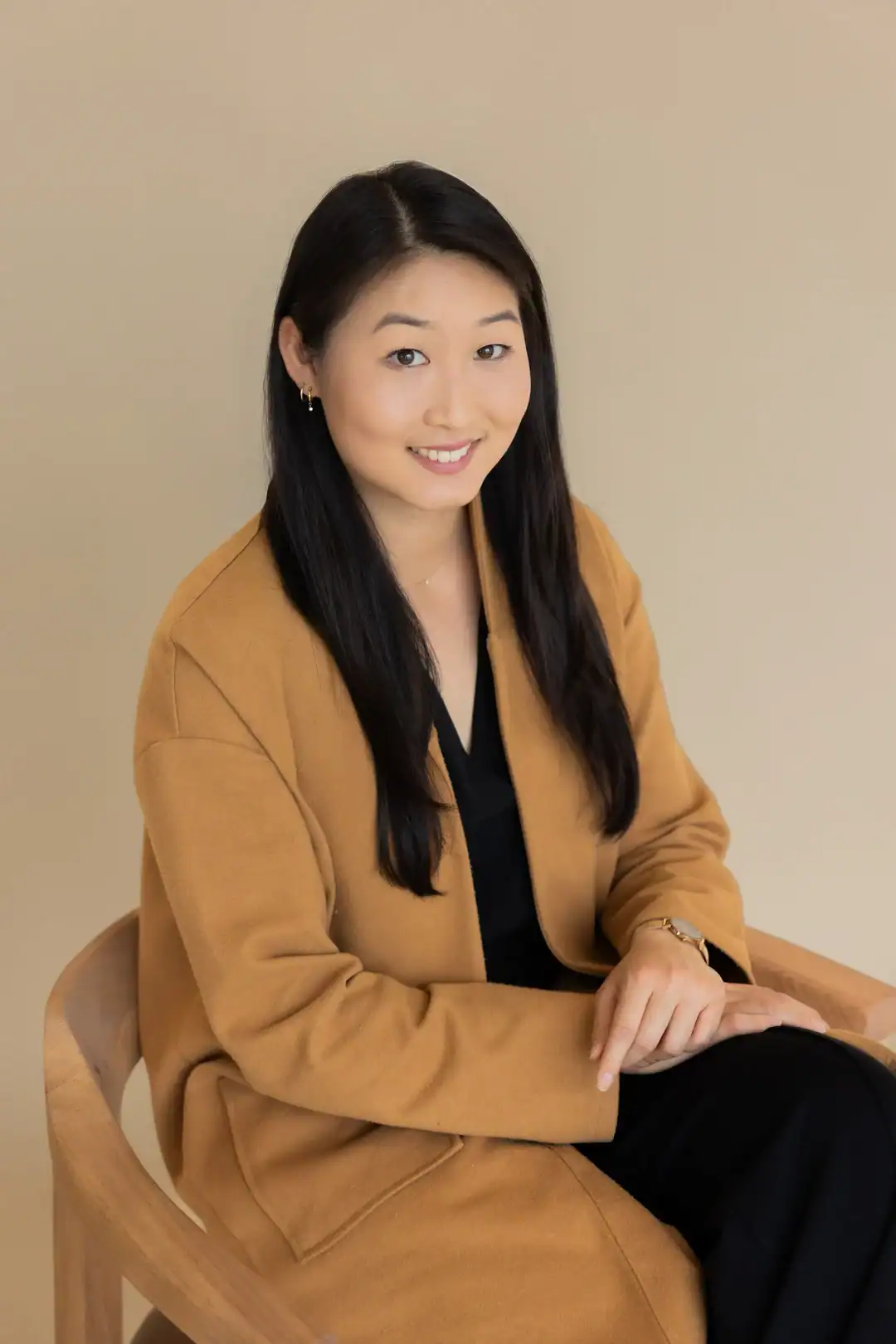 A woman with long black hair wearing a tan coat and black outfit sits on a wooden chair against a beige background, smiling at the camera.