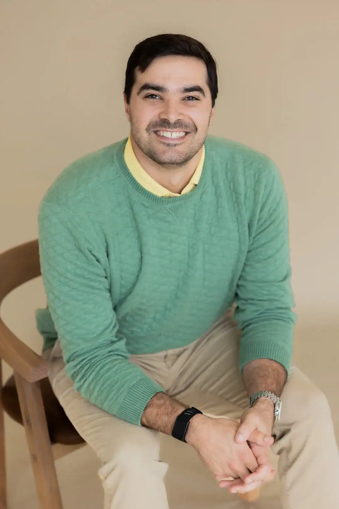Aaron Athanas, a man with dark hair and beard, wearing a green sweater over a yellow shirt and khaki pants, sits on a wooden chair and smiles at the camera against a beige background.
