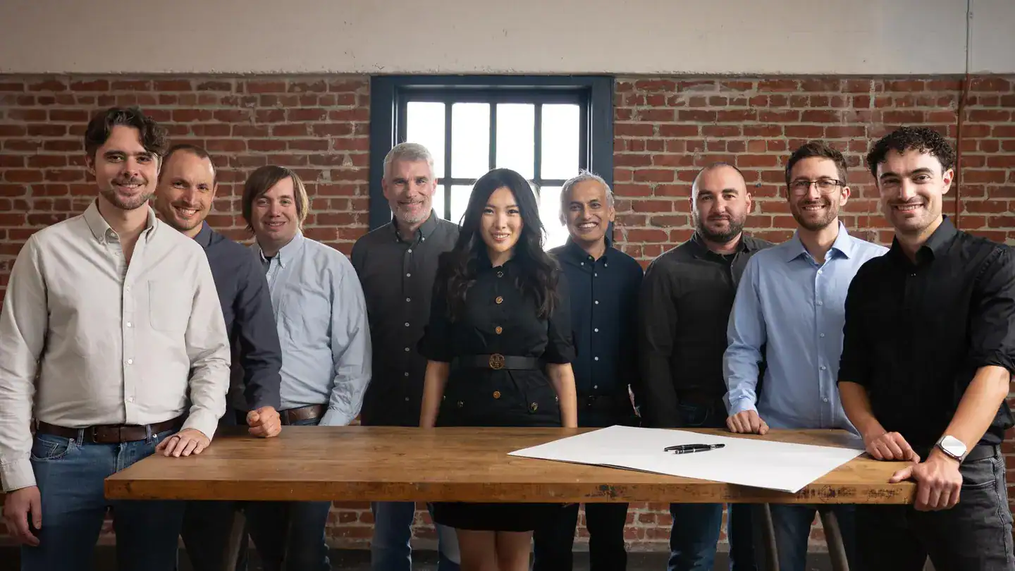 Nine people stand behind a wooden table with paper and pens, in a room with brick walls and a window.