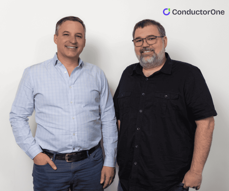 Two men stand side by side and smile at the camera in front of a white wall with the ConductorOne logo in the upper right corner.