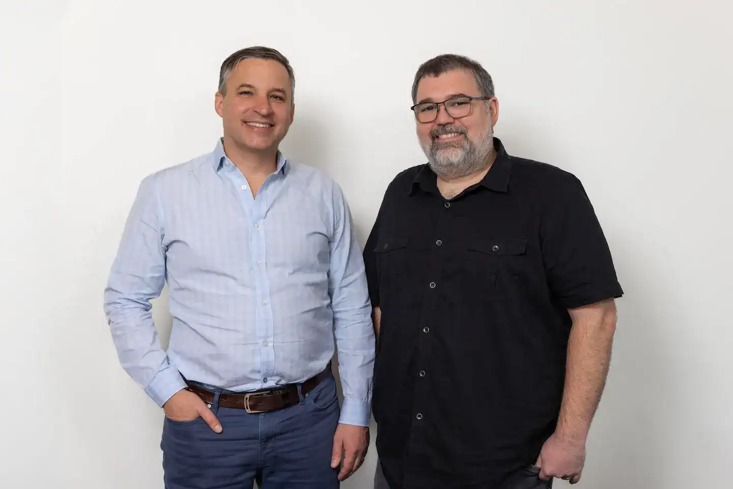 Two men stand side by side against a plain white background, one in a light blue button-up shirt and the other in black, reflecting the modern style of AI employees at a fast-paced startup.