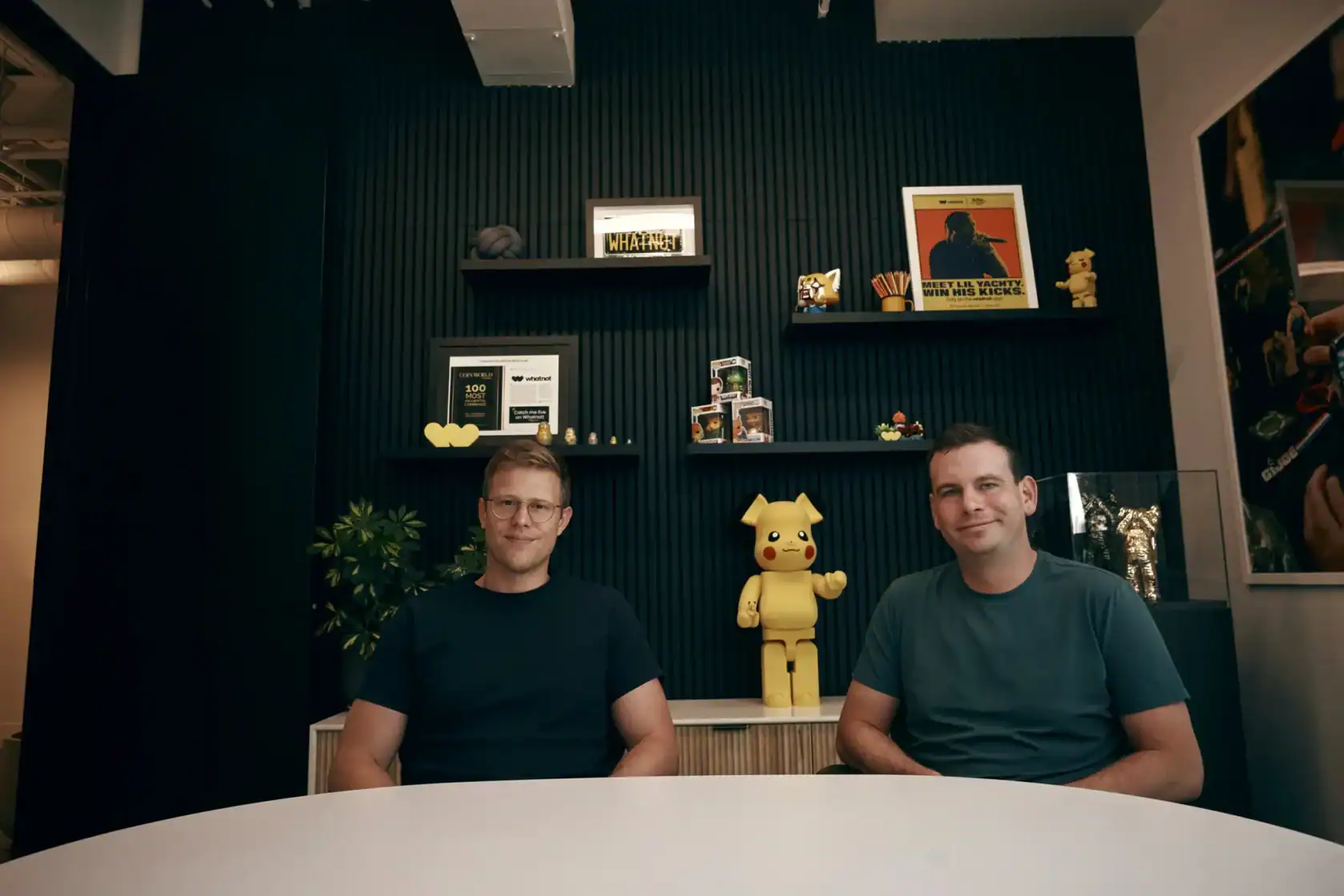 Two men sit at a round table in an office with black walls, shelves displaying awards, books, and Pikachu-themed items behind them.