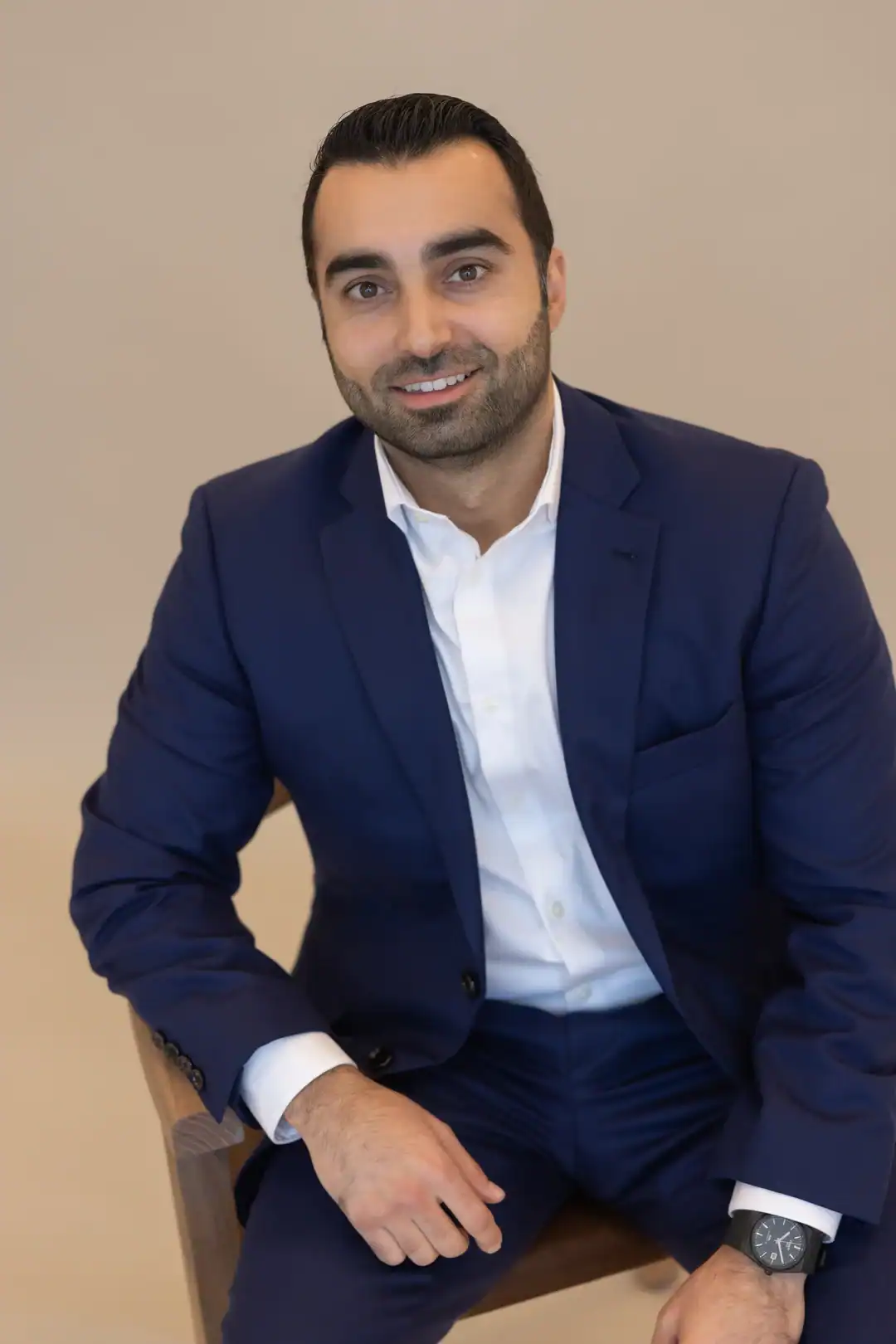 Masoud Adina, dressed in a navy blue suit and white shirt, sits on a chair, smiling at the camera against a plain light-colored background.