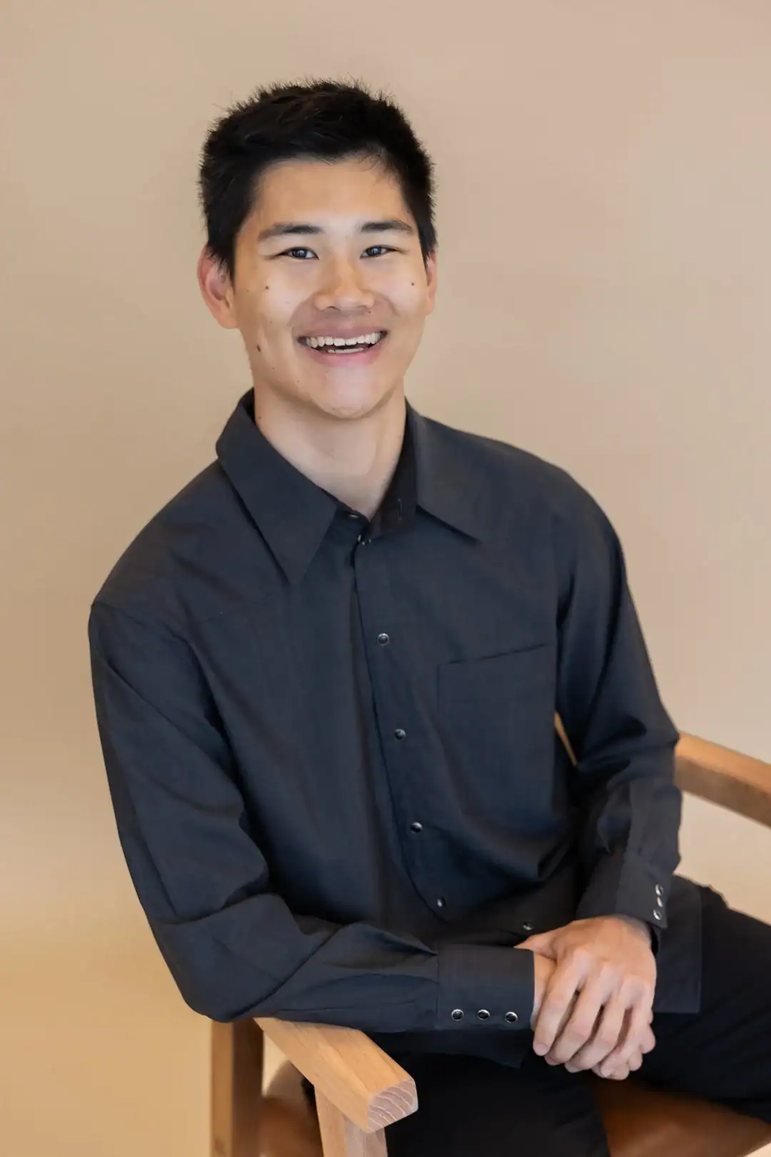 A young man with short black hair, wearing a dark button-up shirt, sits on a wooden chair and smiles at the camera against a beige background.
