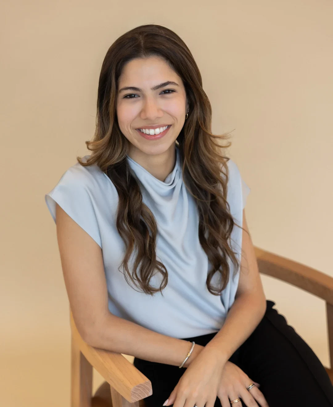 Lauren Arribas, a woman with long brown hair, wearing a light blue blouse and black pants, sits on a wooden chair and smiles at the camera against a beige background.