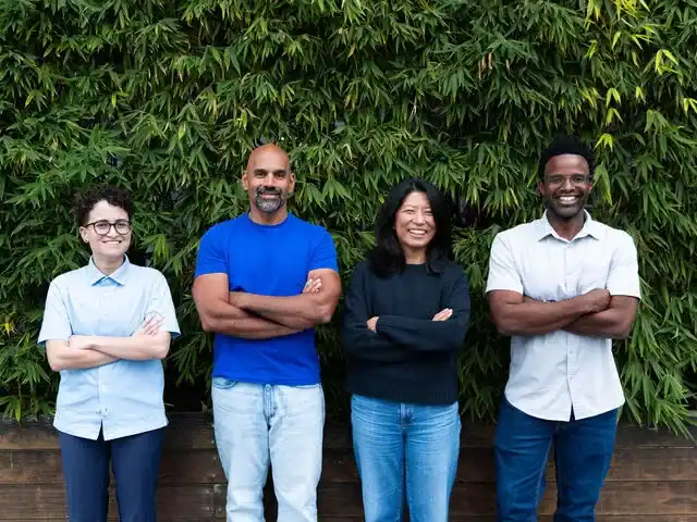 Four people stand side by side in front of green foliage, all with arms crossed and smiling, wearing casual shirts and jeans.