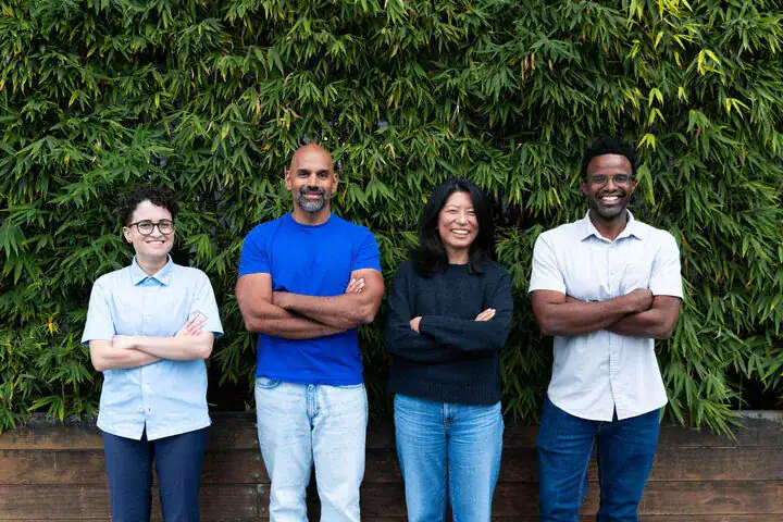 Four people stand side by side in front of green foliage, all with arms crossed and smiling, wearing casual shirts and jeans.