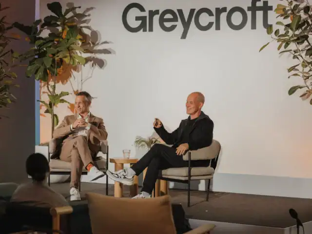 Two men sit on stage in chairs, speaking at an event with a Greycroft sign behind them; plants decorate the stage and an audience member is visible in the foreground, as Brian Robbins discusses his Big Shot backing.