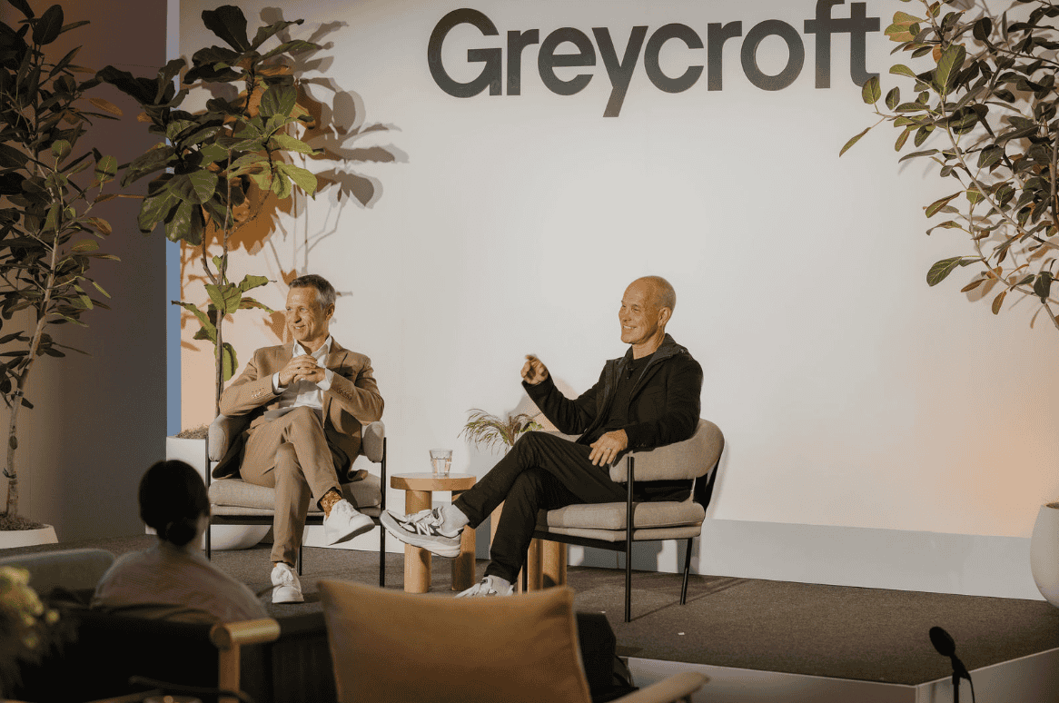 Two men sit on stage in chairs, speaking at an event with a Greycroft sign behind them; plants decorate the stage and an audience member is visible in the foreground, as Brian Robbins discusses his Big Shot backing.
