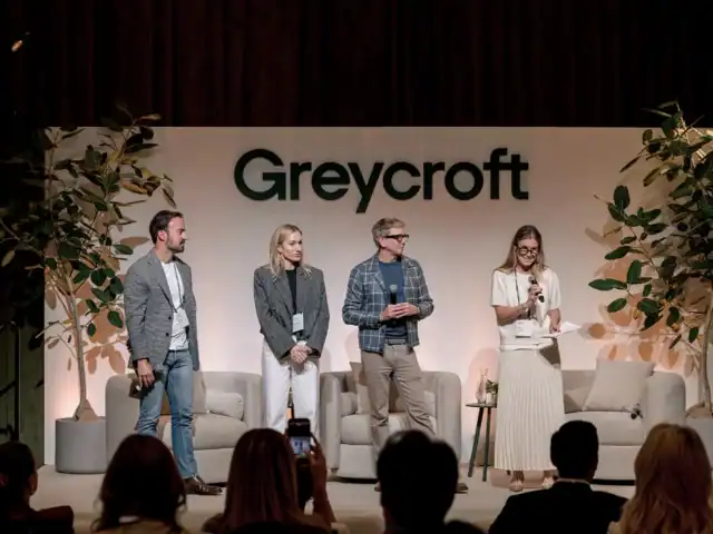 Four people stand on a stage with microphones and notes in front of a Greycroft sign, addressing an audience at an indoor event.