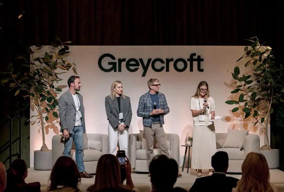 Four people stand on a stage with microphones and notes in front of a Greycroft sign, addressing an audience at an indoor event.