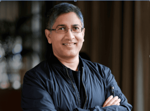 A man with short gray hair and glasses is standing indoors with his arms crossed, smiling at the camera. He is wearing a dark jacket and a black shirt, embodying quiet confidence—much like someone ready to tackle modern challenges such as on-device AI security.