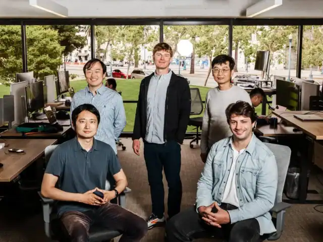Five people pose for a group photo in a modern office with large windows, workstations, and greenery visible outside.