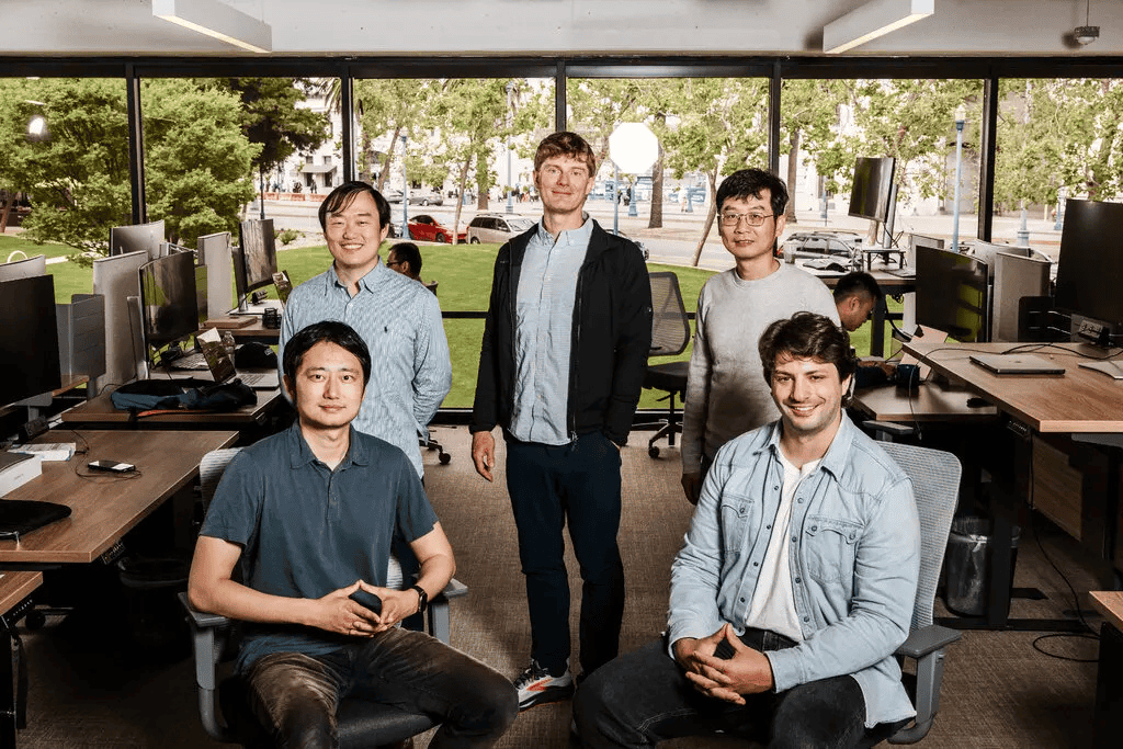 Five people pose for a group photo in a modern office with large windows, workstations, and greenery visible outside.